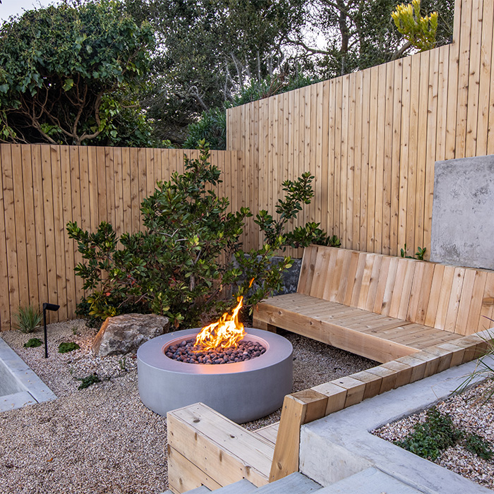 Landscape contruction with a fire pit surrounded by wooden benches, concrete structures, wooden fences and gravel paths.